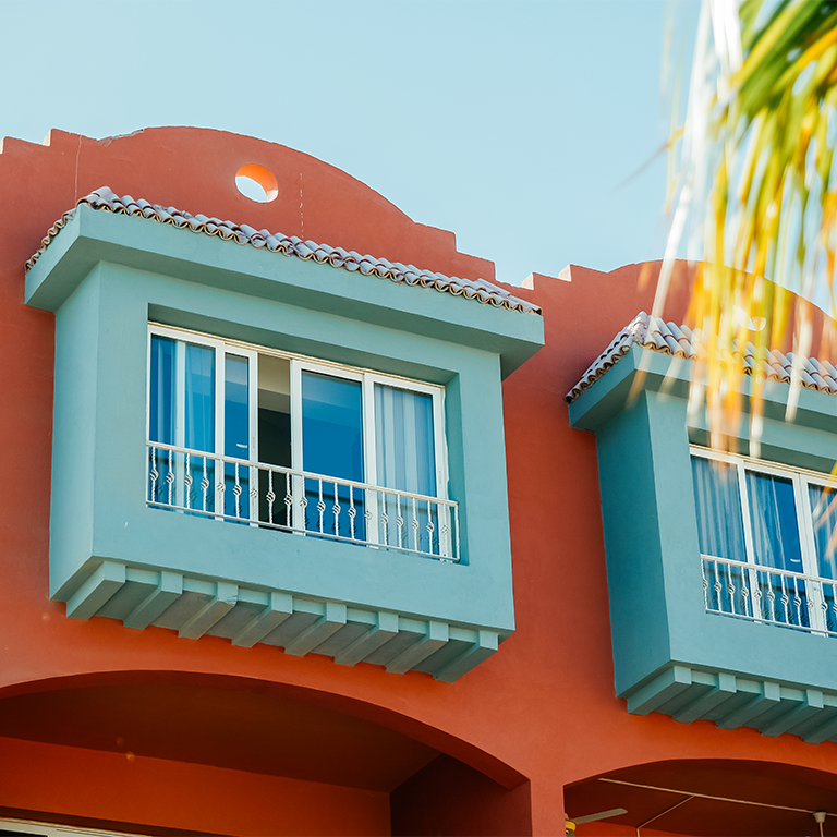 Terracotta coastal holiday home abroad with turquoise bay windows and sunlit palm fronds.