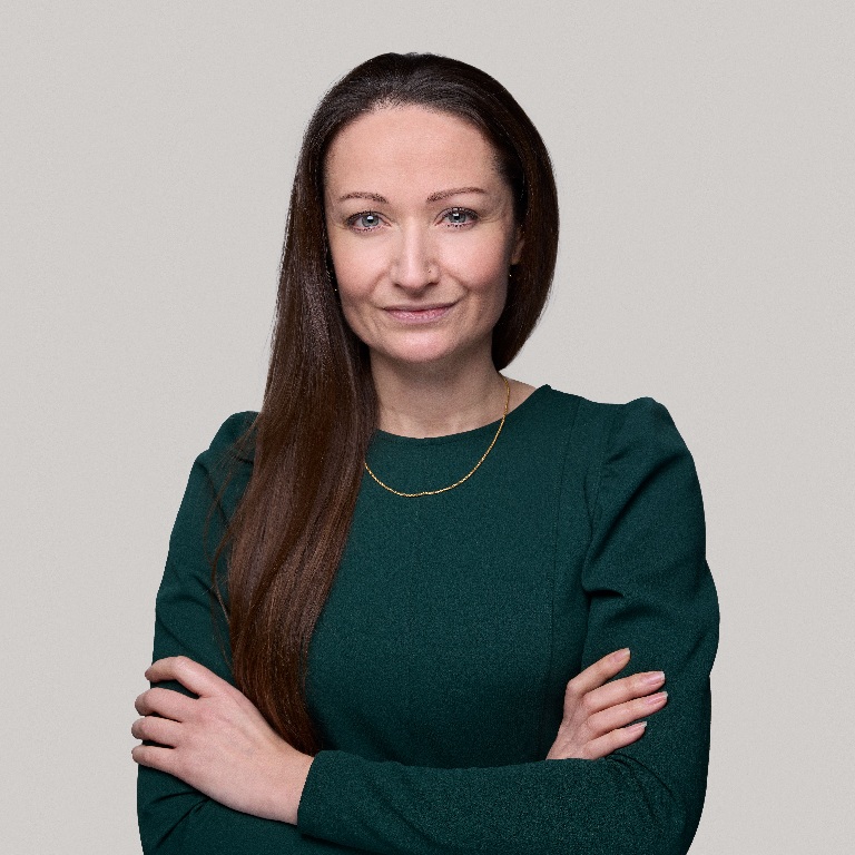 A woman with long dark hair standing with arms crossed, wearing a dark green long-sleeve top and a thin gold necklace against a light gray background.
