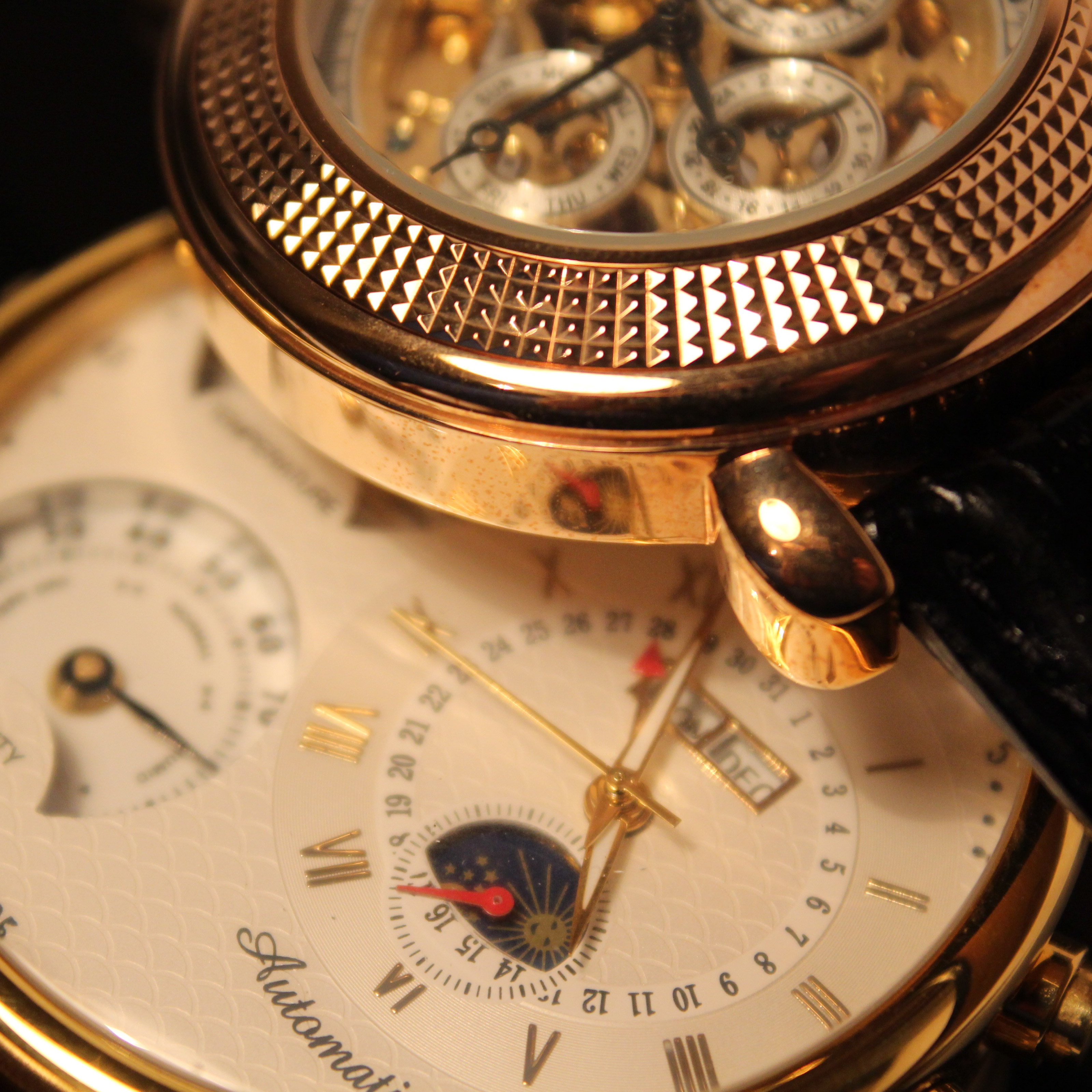 Close-up of two gold luxury wristwatches with detailed dials, visible hands, moonphase and subdials on a white face and black leather strap for a Bonhams interview.