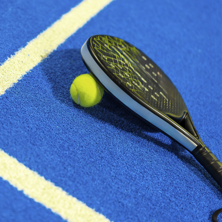 Close-up of a padel racket and yellow ball resting on a blue padel court with white boundary lines, highlighting the textured court surface