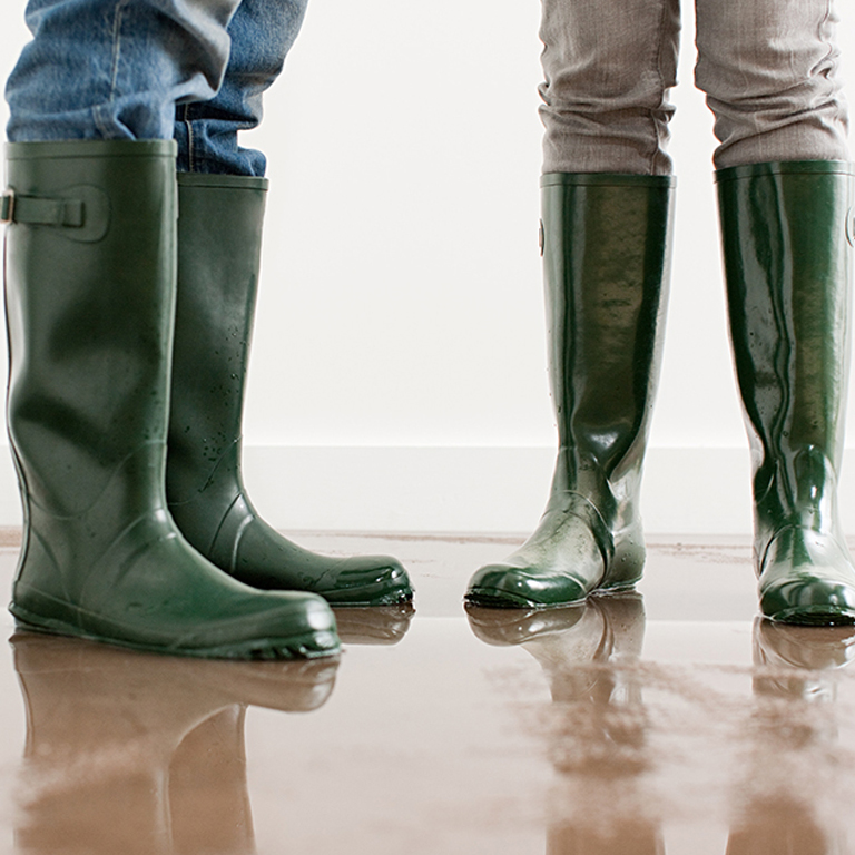 Two people in green rubber boots stand on a wet hardwood floor with pooled water from water escape damage indoors